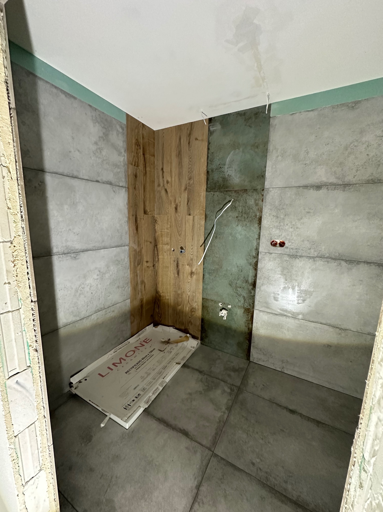 Wnętrze łazienki w trakcie prac wykończeniowych. Widoczne płytki imitujące beton i drewno, wystające kable oraz opakowanie po płytkach na podłodze.