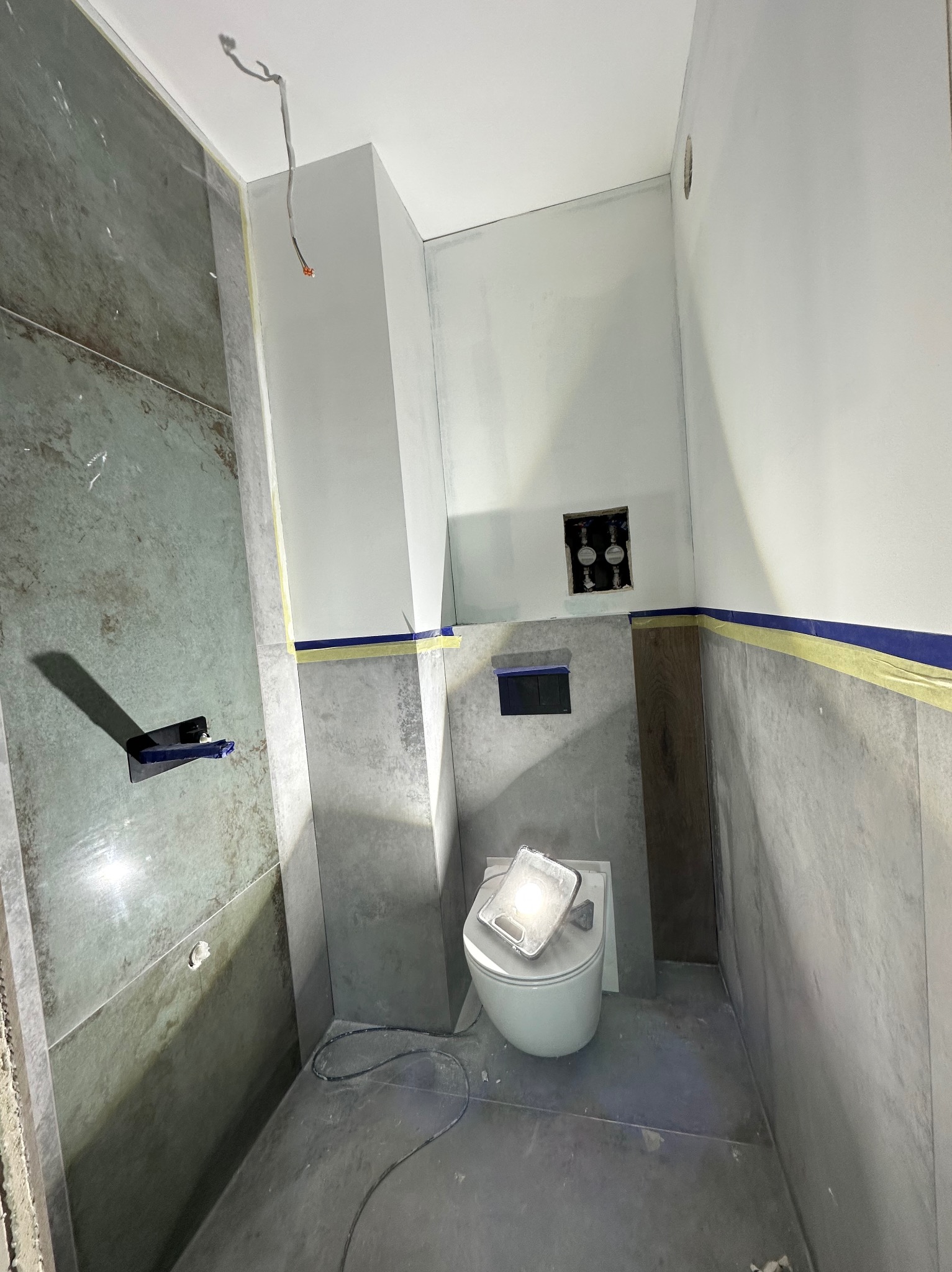 Wnętrze łazienki w trakcie prac wykończeniowych: częściowo położone płytki imitujące beton, przygotowane ściany do malowania, wisząca toaleta z lampą roboczą.