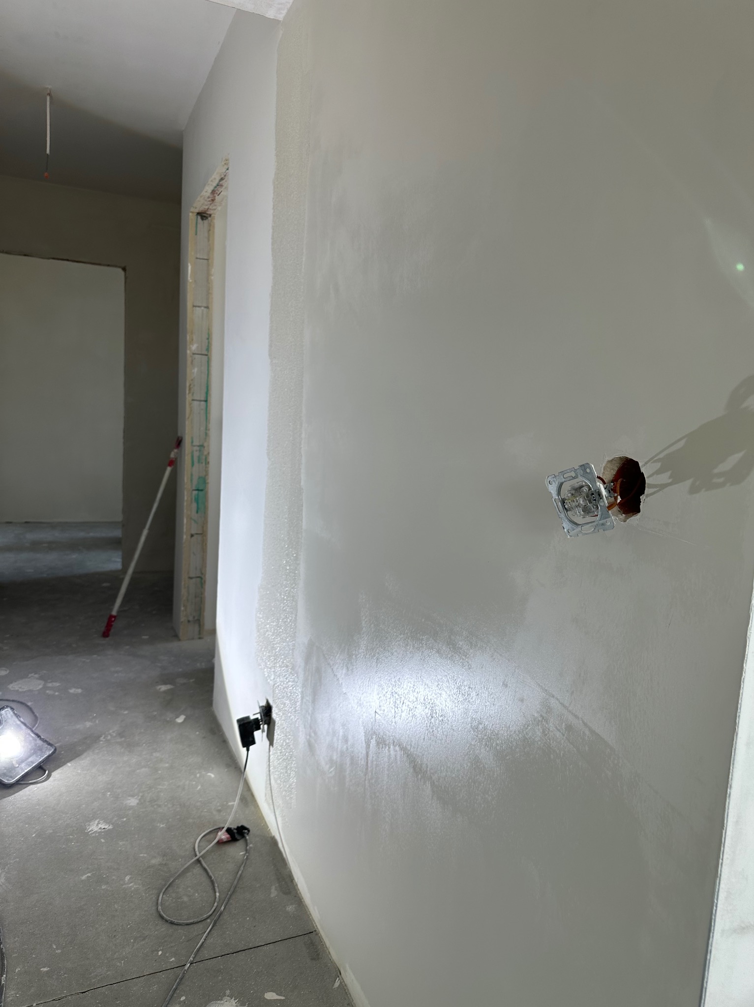 Świeżo wyszpachlowana ściana z widocznym gniazdkiem elektrycznym, w tle wałek malarski i lampa budowlana. Widoczne ślady szpachlowania.