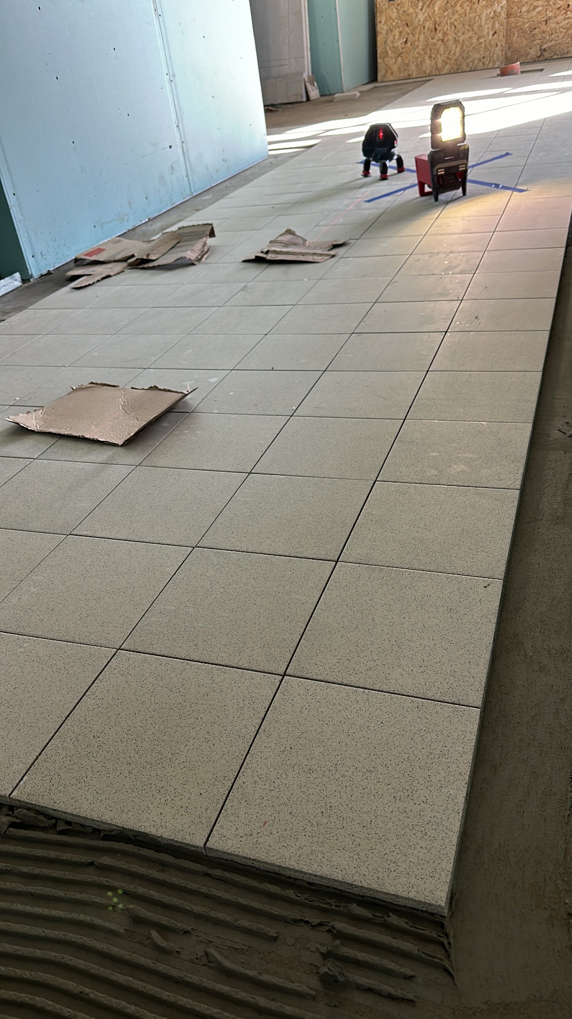 Ułożone kwadratowe płytki podłogowe w trakcie prac wykończeniowych, z widocznym laserowym niwelatorem i fragmentami kartonu ochronnego. Szare płytki z delikatnym wzorem.