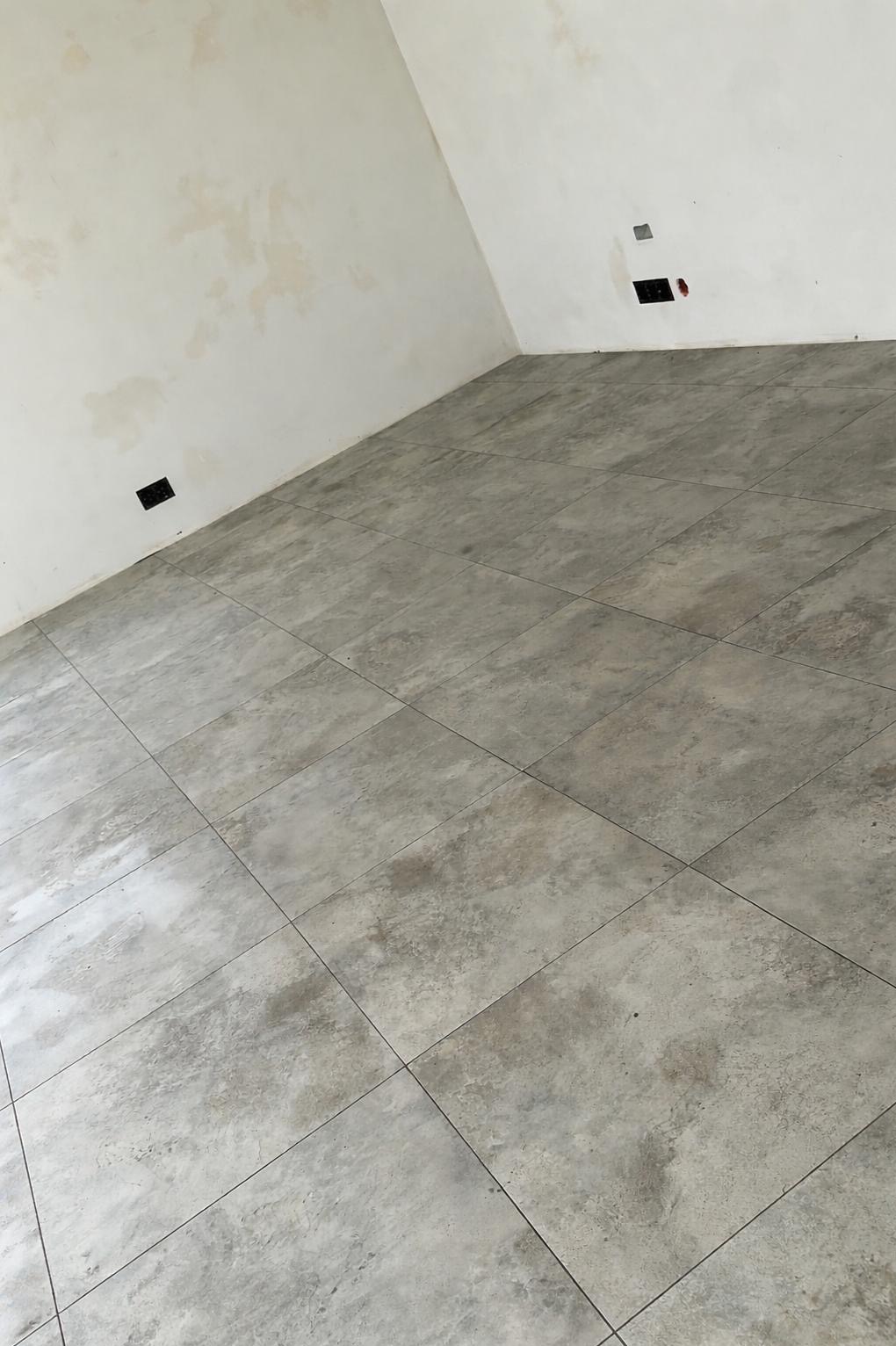 Ukończona podłoga z szarych, kwadratowych płytek w nowym pomieszczeniu z surowymi ścianami i widocznymi otworami na gniazdka elektryczne.