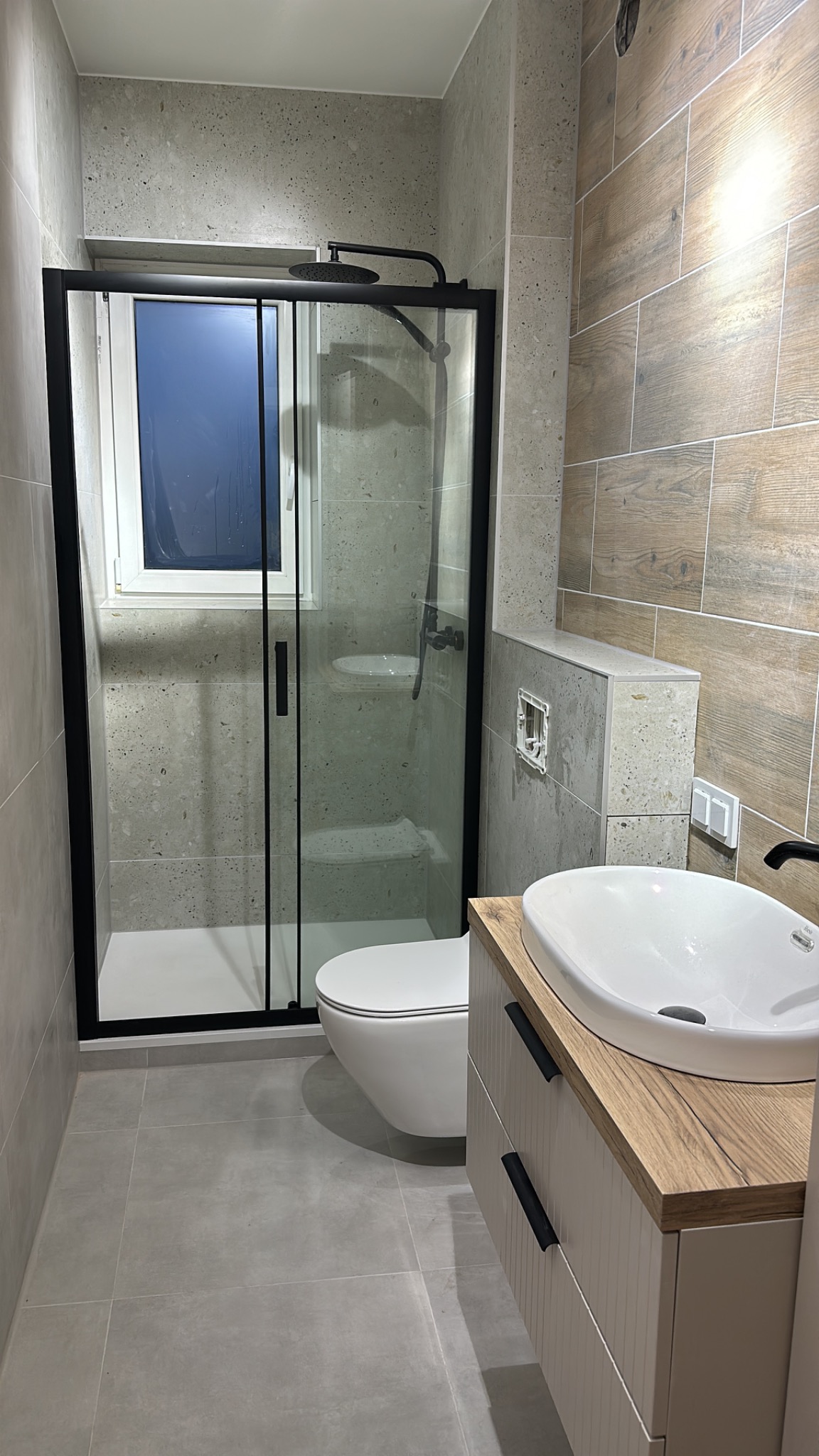Nowoczesna łazienka z kabiną prysznicową typu walk-in, umywalką nablatową na szafce z drewnianym blatem i płytkami imitującymi drewno na ścianie.