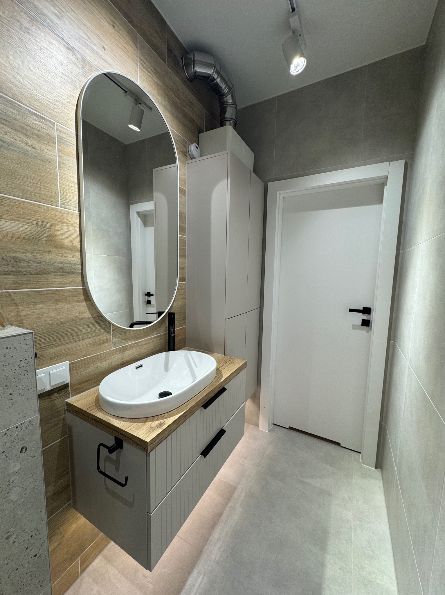 Nowoczesna łazienka z owalnym lustrem, białą umywalką nablatową na szafce z szufladami i drewnianym blatem. Beżowe płytki imitujące drewno na ścianie.