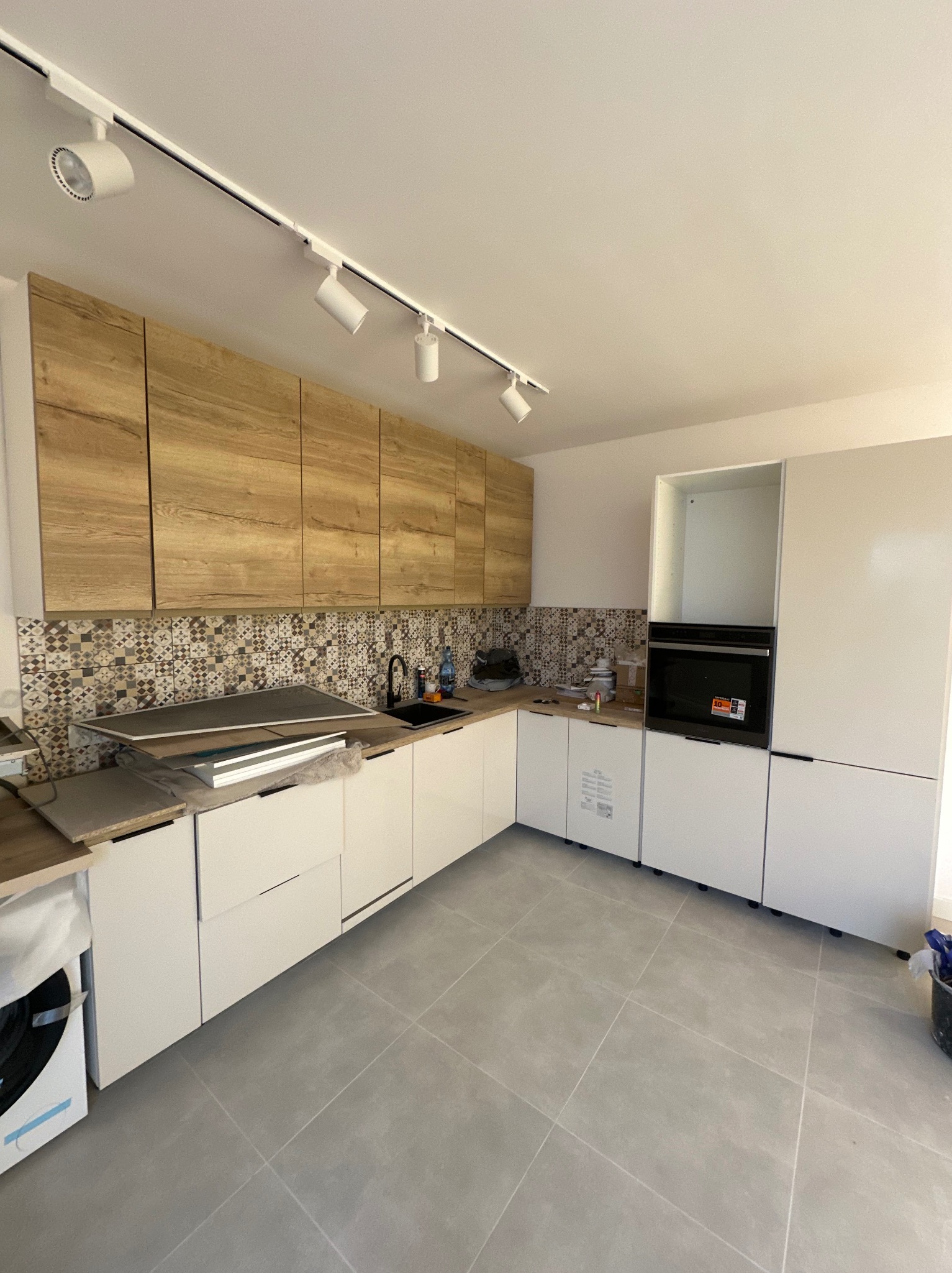 Jasna kuchnia w trakcie montażu: białe szafki dolne, drewnopodobne górne, mozaika nad blatem. Widoczne płyty do montażu i szare płytki podłogowe.
