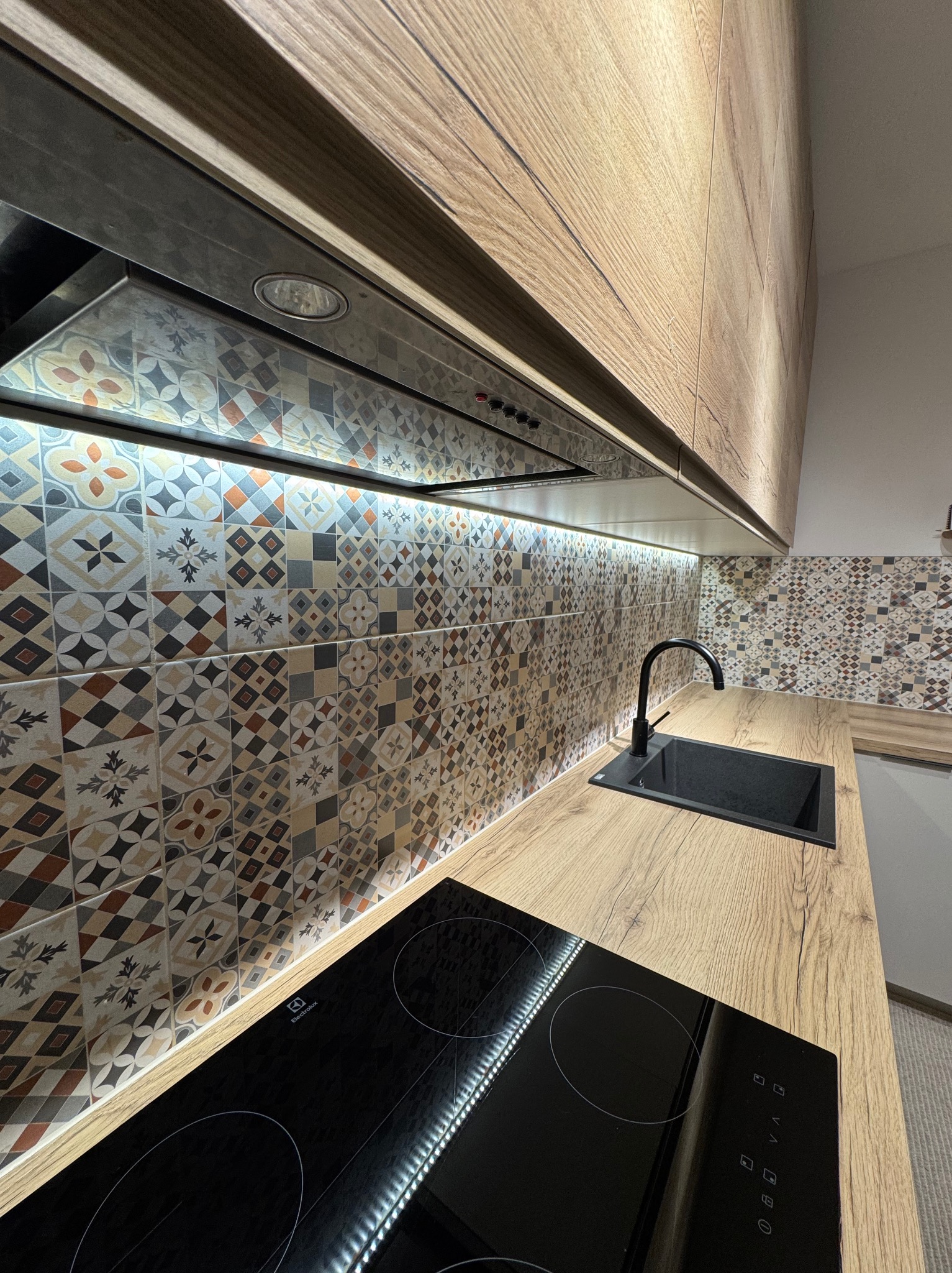 Nowoczesna kuchnia z drewnianymi szafkami, mozaiką na ścianie i czarnym zlewem. Oświetlenie LED pod szafkami dodaje blasku. Widoczna płyta indukcyjna.