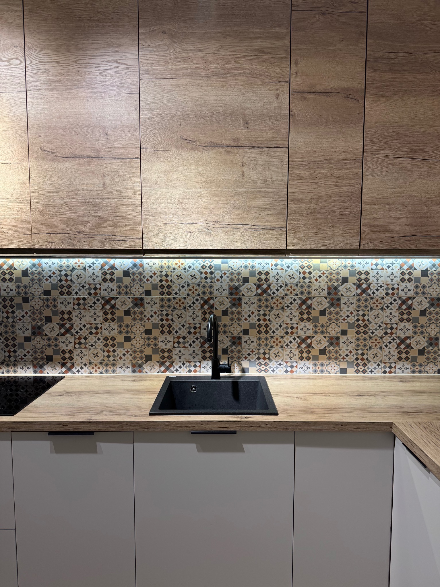 Nowoczesna kuchnia z drewnianymi szafkami, mozaiką nad blatem i czarnym zlewem. Oświetlenie LED podkreśla wzór płytek. Minimalistyczny design.