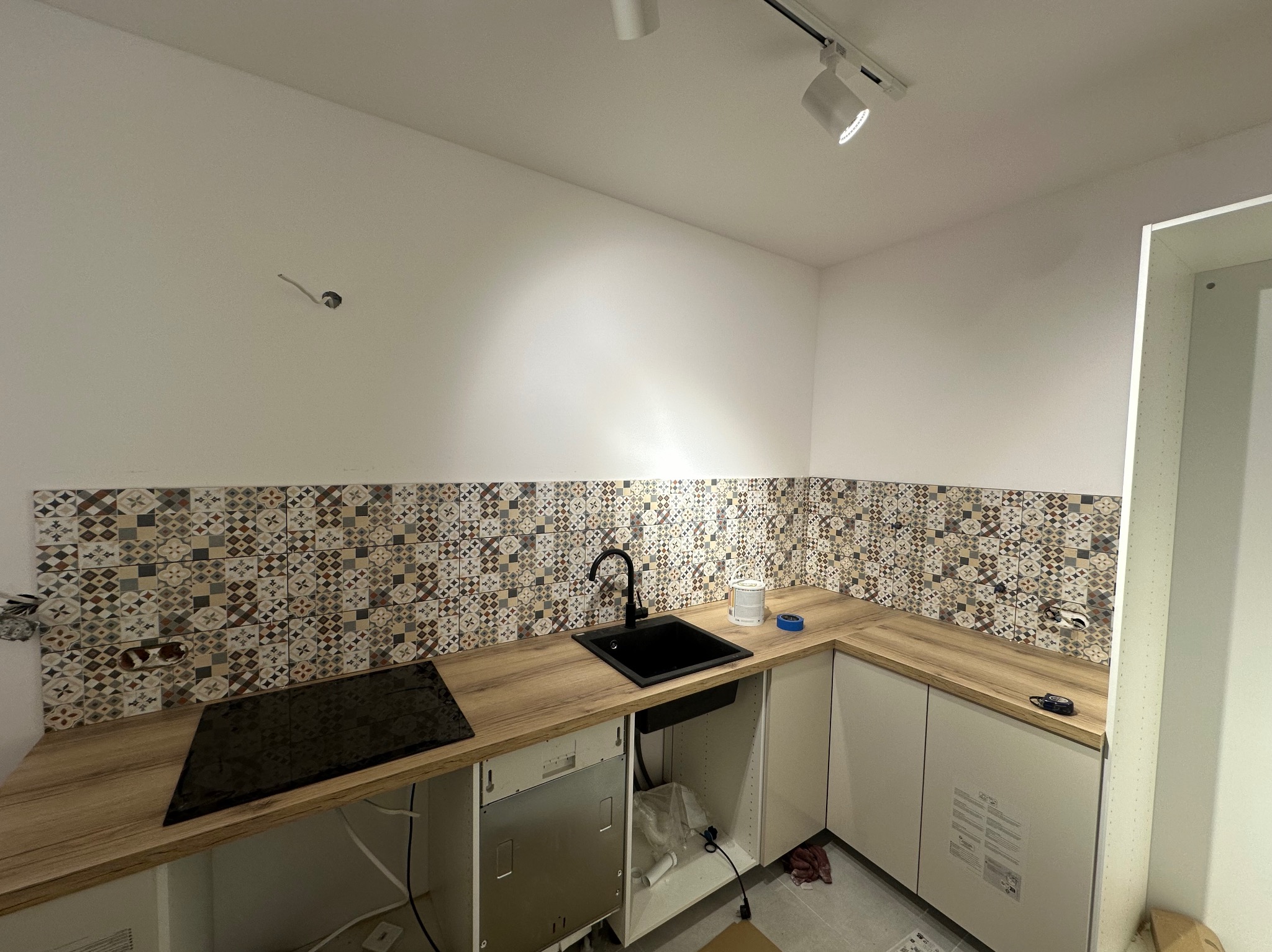 Montaż płytek dekoracyjnych nad blatem w kuchni. Widoczny czarny zlew, płyta indukcyjna i białe szafki. Oświetlenie punktowe na suficie. Etap prac wykończeniowych.