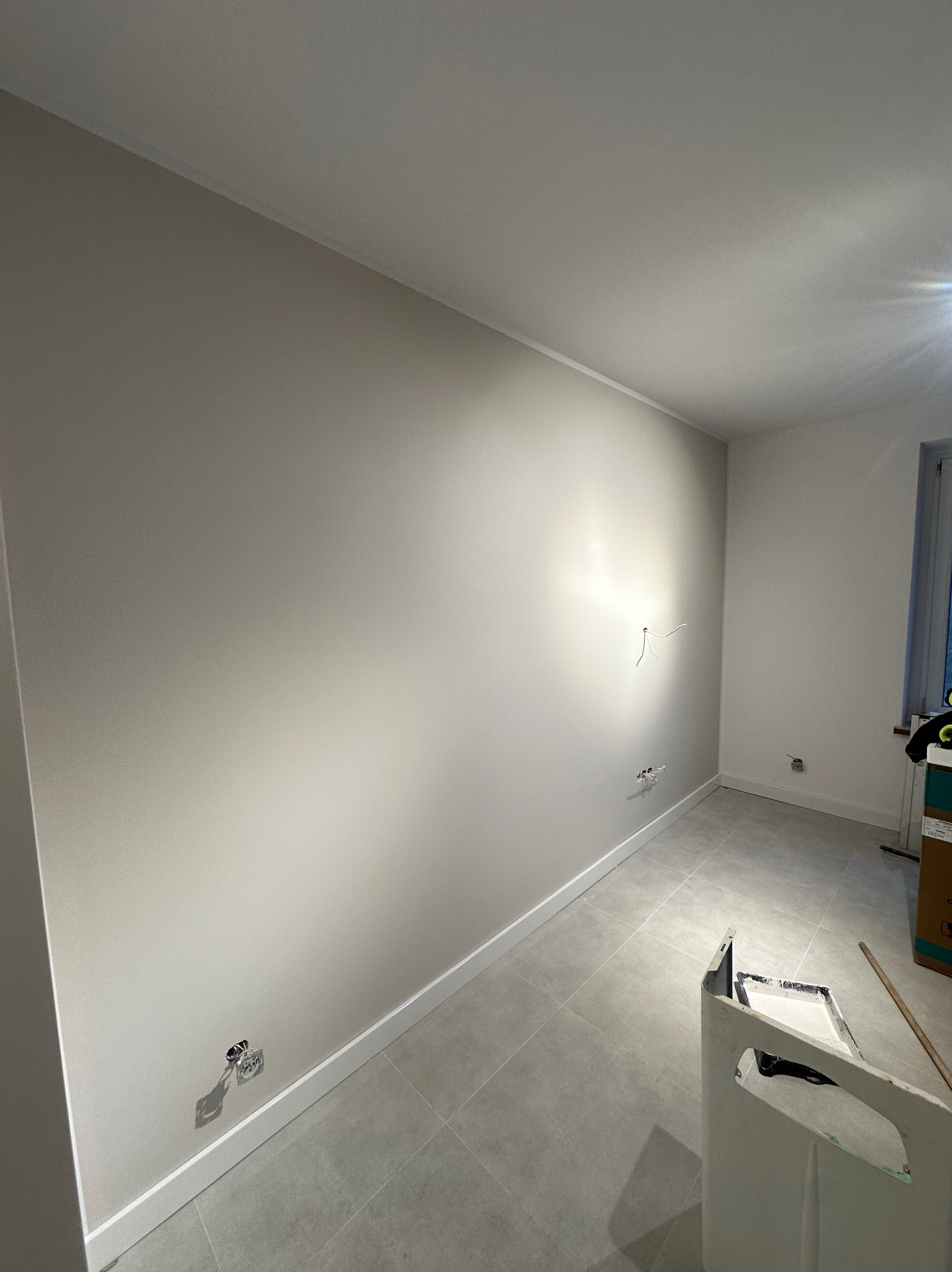 Jasne wnętrze po malowaniu ścian na beżowo, z widocznymi gniazdkami elektrycznymi i listwami przypodłogowymi. Na podłodze szare płytki. Widoczny kuwet z farbą.