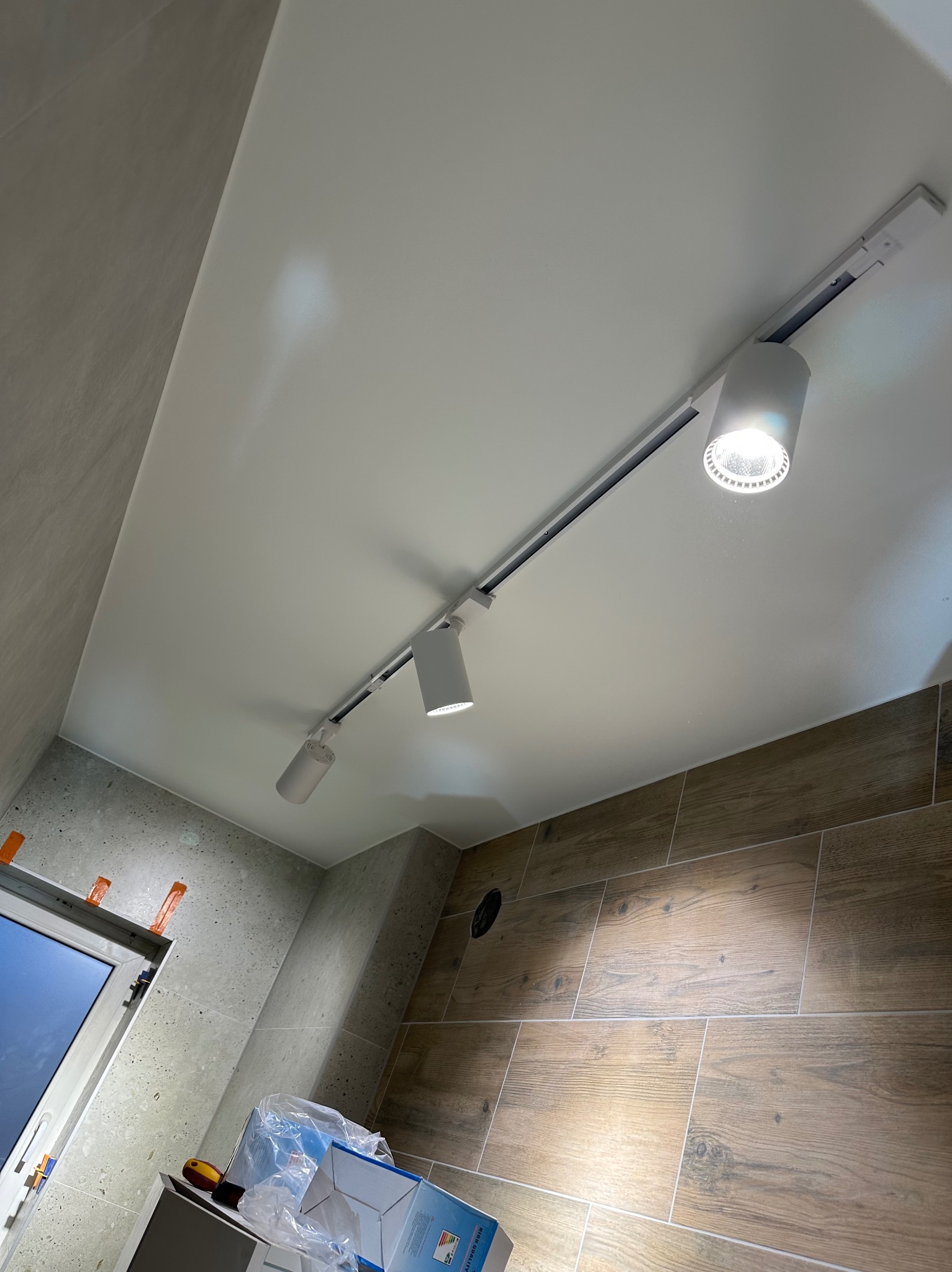 Nowoczesne oświetlenie szynowe z trzema reflektorami w wykończonej łazience z płytkami imitującymi drewno i szarym gresem. Widok pod kątem.