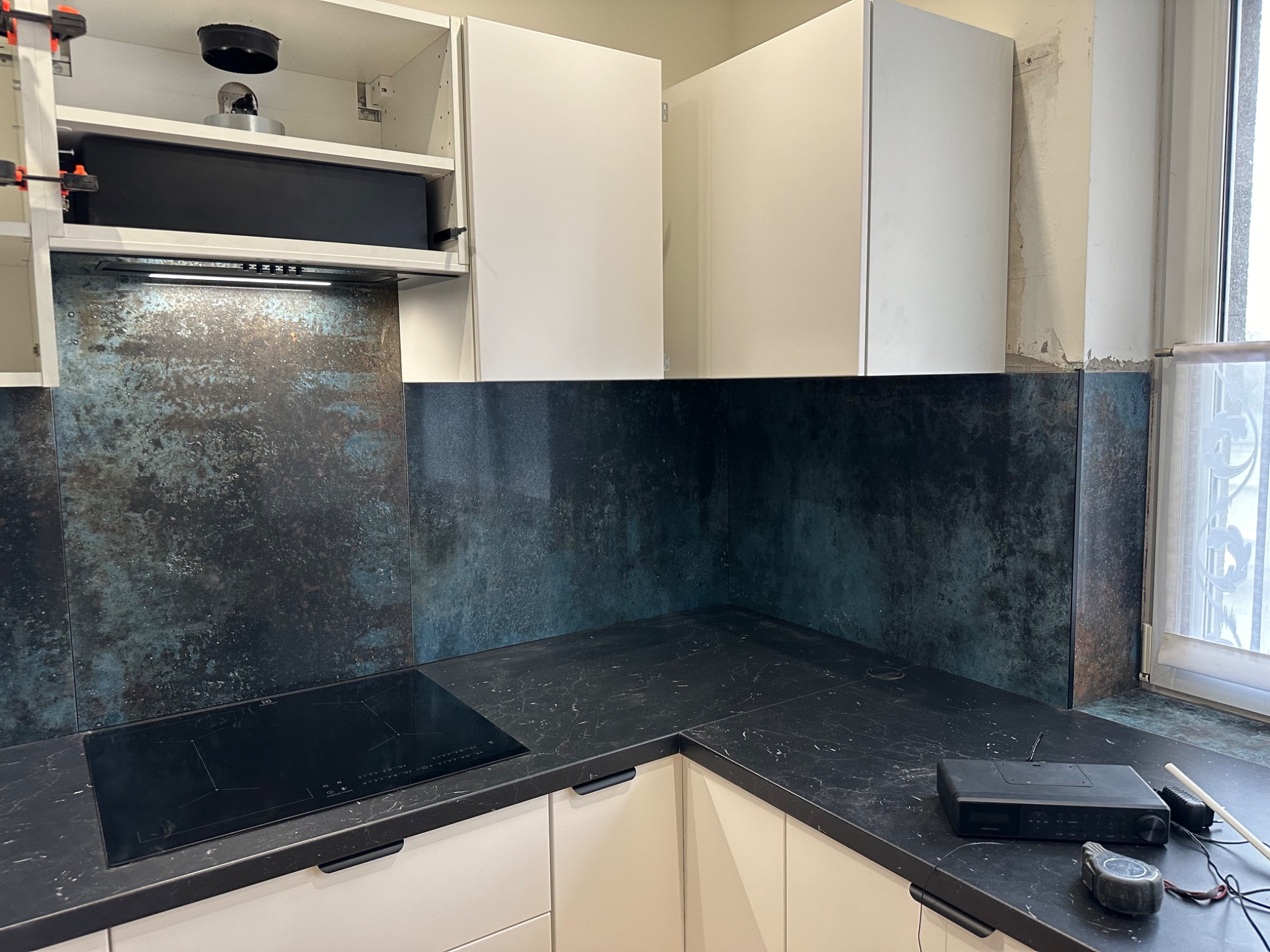 Nowoczesna kuchnia w trakcie montażu: ciemny blat, indukcja, metaliczne panele ścienne. Widoczne narzędzia i niedokończone wykończenie przy oknie. Białe szafki.