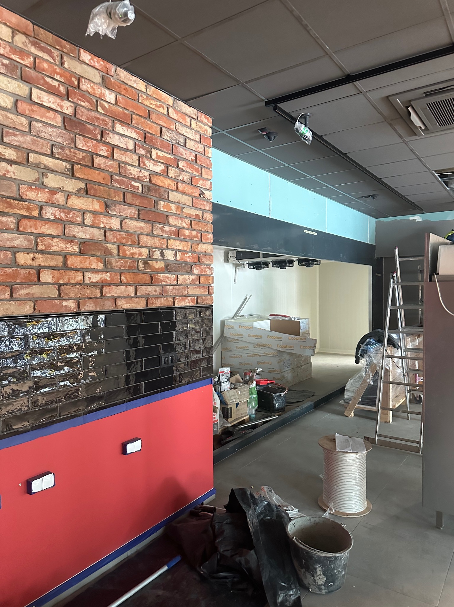 Wnętrze w trakcie prac wykończeniowych: ściana z cegły i czarnych płytek, czerwona dolna część ściany, widoczne materiały budowlane i drabina. Oświetlenie sufitowe.