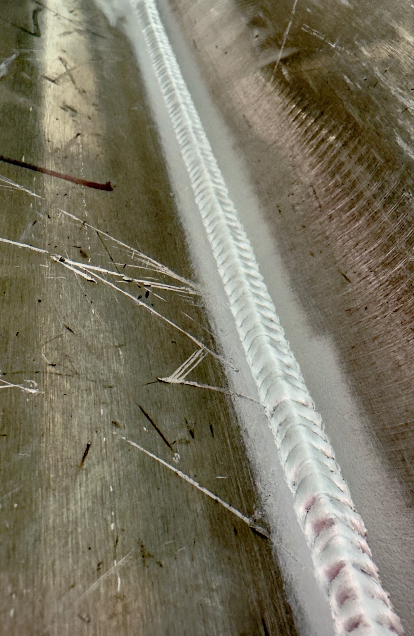 Szlifowana powierzchnia metalu ze świeżym, równym spoiwem spawalniczym pośrodku, widoczne drobne odpryski i rysy. Ukazany detal wykonanej pracy.