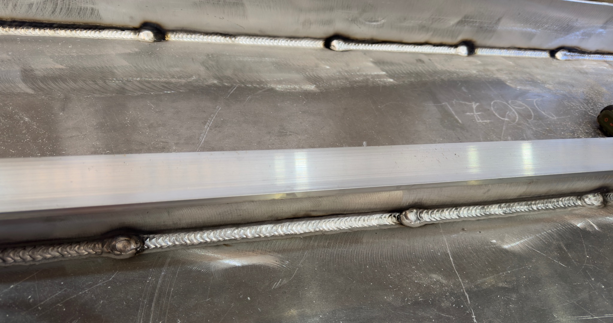 Szlifowana powierzchnia metalowa z widocznymi spawami i profilem aluminiowym. Widoczne ślady po obróbce i numer zapisany na powierzchni.