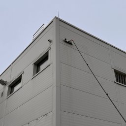 GRUPA ECS DOMINIK JAROCKI - Mycie okien na wysokości budynku przemysłowego za pomocą długiej lancy. Widoczne okna, elewacja z paneli, kamery i oświetlenie zewnętrzne. Ujęcie z dołu.