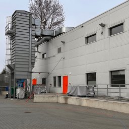 GRUPA ECS DOMINIK JAROCKI - Szary budynek przemysłowy z systemem wentylacji Nestro, widoczne worki i pojemniki na odpady, teren wyłożony kostką brukową, pochmurne niebo.