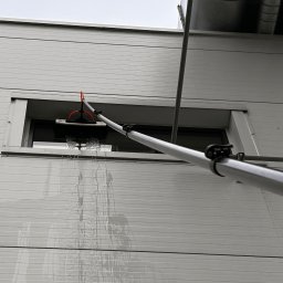 GRUPA ECS DOMINIK JAROCKI - Mycie okna na wysokości za pomocą teleskopowego kija z myjką. Woda spływa po elewacji. Widoczna szara elewacja budynku z poziomymi panelami.