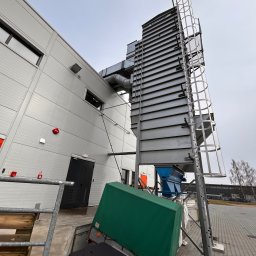 GRUPA ECS DOMINIK JAROCKI - Zewnętrzny system wentylacji przemysłowej z metalową konstrukcją i drabinką, umieszczony przy szarej hali z widocznymi elementami instalacji i zielonym kontenerem.