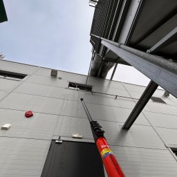 CleanTech.pl - Grupa ECS - Mycie okien na wysokości w budynku przemysłowym za pomocą teleskopowej lancy. Widoczne ślady spływającej wody po czyszczeniu. Czerwona lanca z oznaczeniami bezpieczeństwa.