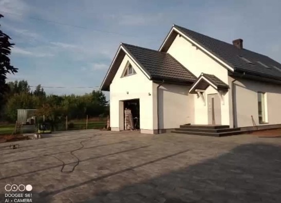 Nowoczesny dom z szarą kostką brukową na podjeździe. Widoczne schody wejściowe i garaż. Tło stanowią drzewa i błękitne niebo. Ujęcie z niskiej perspektywy.