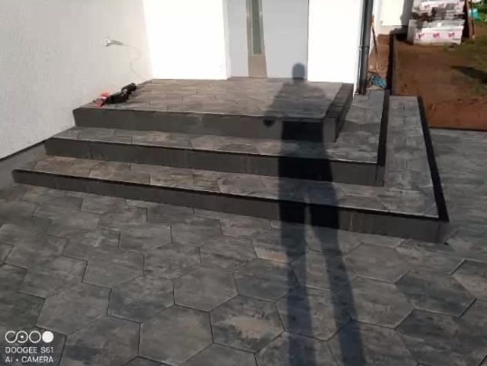 Schody wejściowe i chodnik z szarej, heksagonalnej kostki brukowej. Widoczne cienie osoby fotografującej. W tle fragment budynku i trawnika.