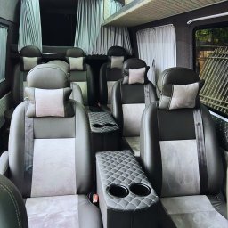 POLONIA BUS Busy do Holandii, Niemiec, Belgii, Szwajcarii, Włoch i Austrii - Luksusowe wnętrze busa z szarymi skórzanymi fotelami, pikowanymi podłokietnikami z uchwytami na napoje i zasłonami w oknach, tworzące komfortową przestrzeń podróżną.