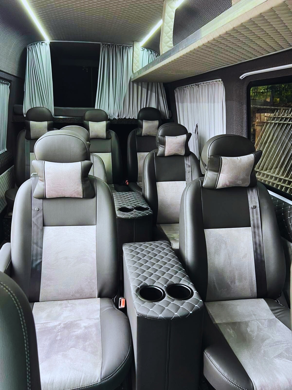 Luksusowe wnętrze busa z szarymi skórzanymi fotelami, pikowanymi podłokietnikami z uchwytami na napoje i zasłonami w oknach, tworzące komfortową przestrzeń podróżną.