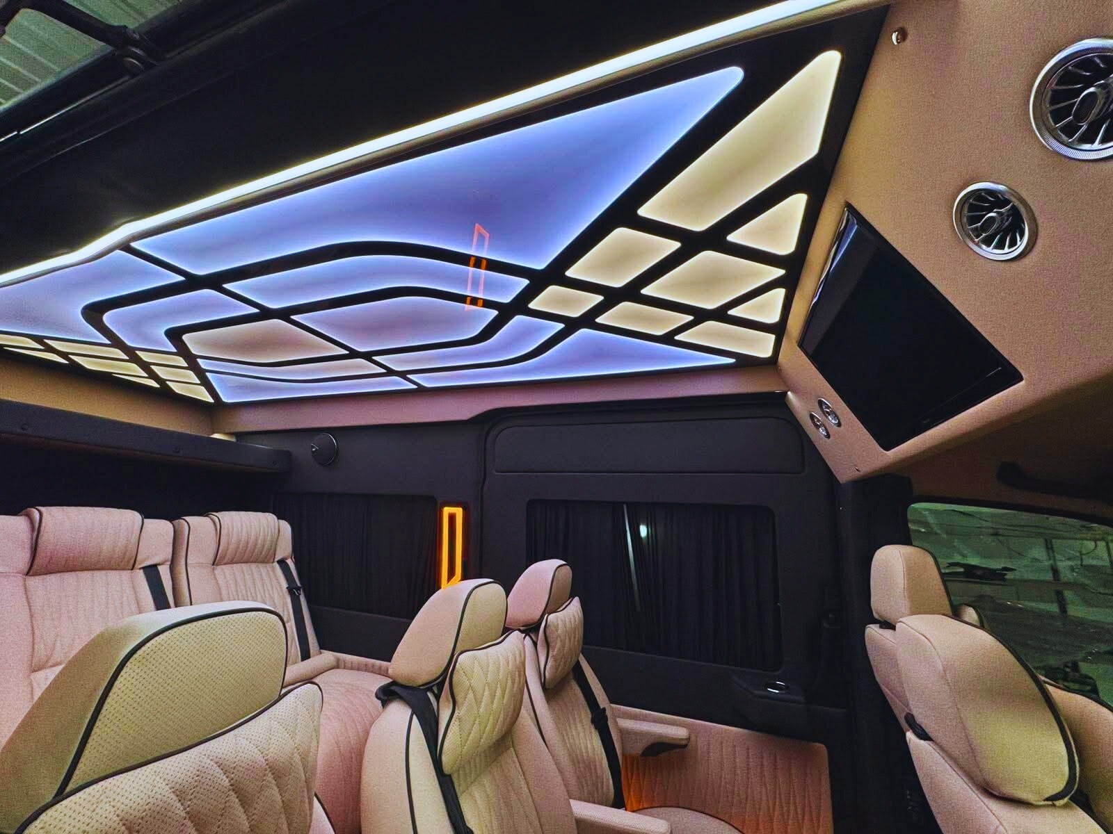 Luksusowe wnętrze busa z beżowymi skórzanymi fotelami, podświetlanym sufitem z geometrycznym wzorem i zamontowanym telewizorem. Detale wykończenia w kolorze czarnym.