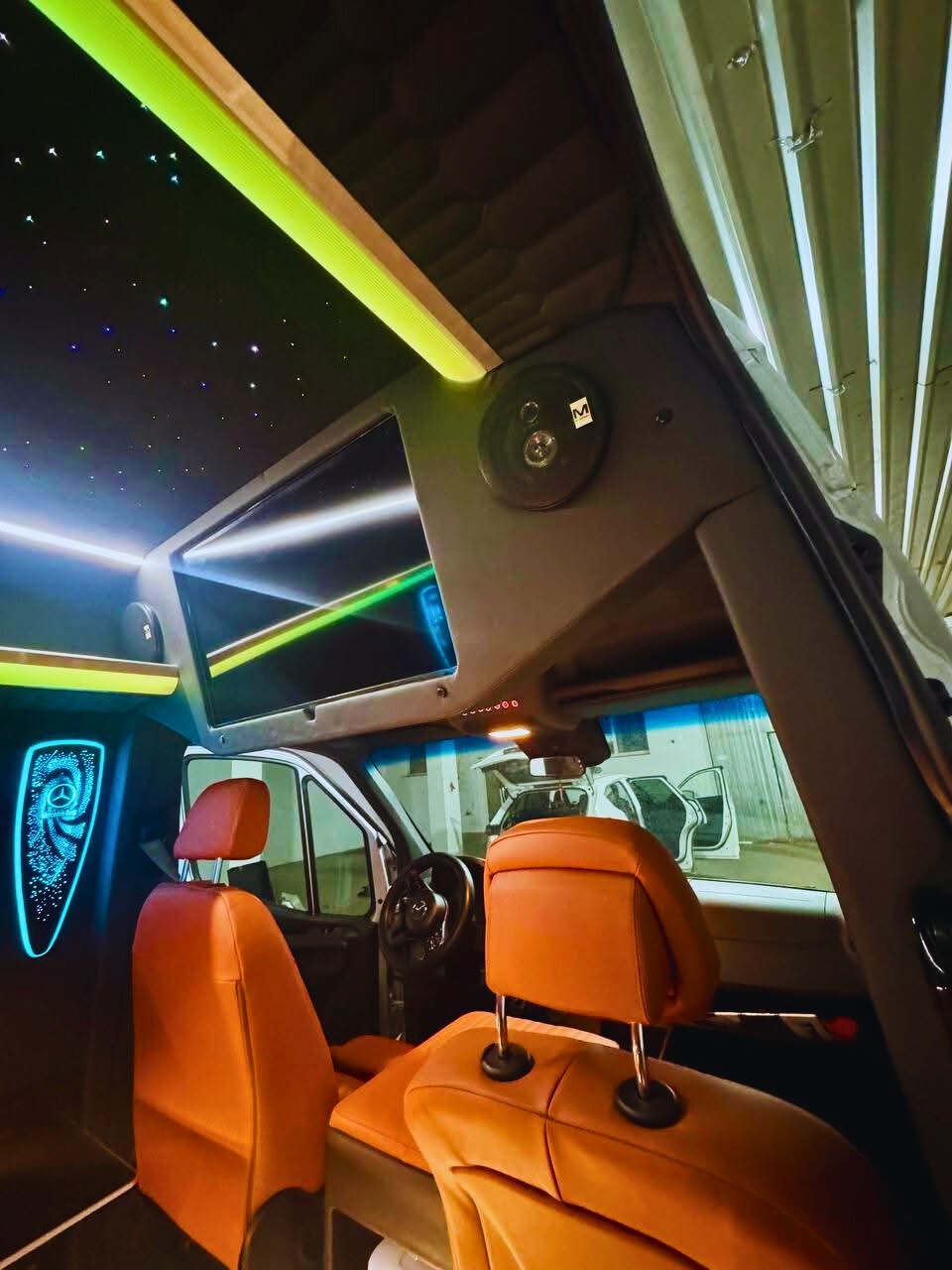 Luksusowe wnętrze busa z pomarańczowymi skórzanymi fotelami, oświetleniem LED i panelem multimedialnym. Widok z perspektywy pasażera na kabinę kierowcy.