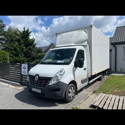 DanLogistics - Biały bus dostawczy Renault Master z zabudową kontenerową, zaparkowany na brukowanej ulicy przed posesją z zielenią i ogrodzeniem. Widoczna tablica rejestracyjna.