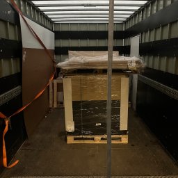 DanLogistics - Paleta z towarem zabezpieczona folią i pasami wewnątrz przestrzeni ładunkowej busa. Widoczne elementy mocujące i podłoga z blachy ryflowanej.