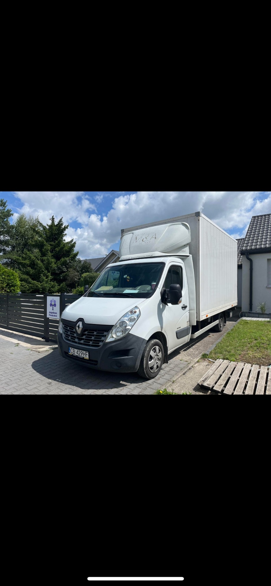 Biały bus dostawczy Renault Master z zabudową kontenerową, zaparkowany na brukowanej ulicy przed posesją z zielenią i ogrodzeniem. Widoczna tablica rejestracyjna.