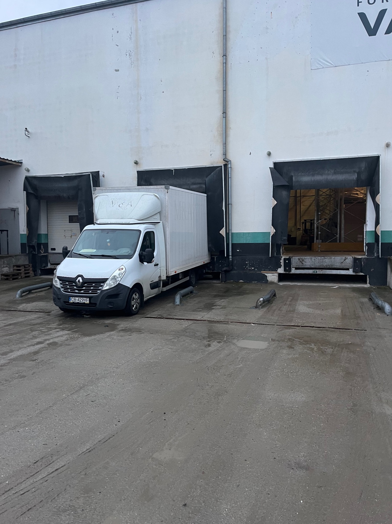 Biały bus dostawczy Renault Master zaparkowany przed rampą magazynową z otwartymi wrotami. Widoczny numer rejestracyjny pojazdu i fragment logo na nadbudowie.