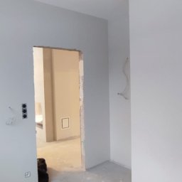 4 Kąty Jarosław Ogeniewski - Wnętrze w trakcie prac wykończeniowych: świeżo pomalowane ściany na szaro, surowa podłoga, widoczne kable i gniazdka. W tle otwarte przejście do innego pomieszczenia.