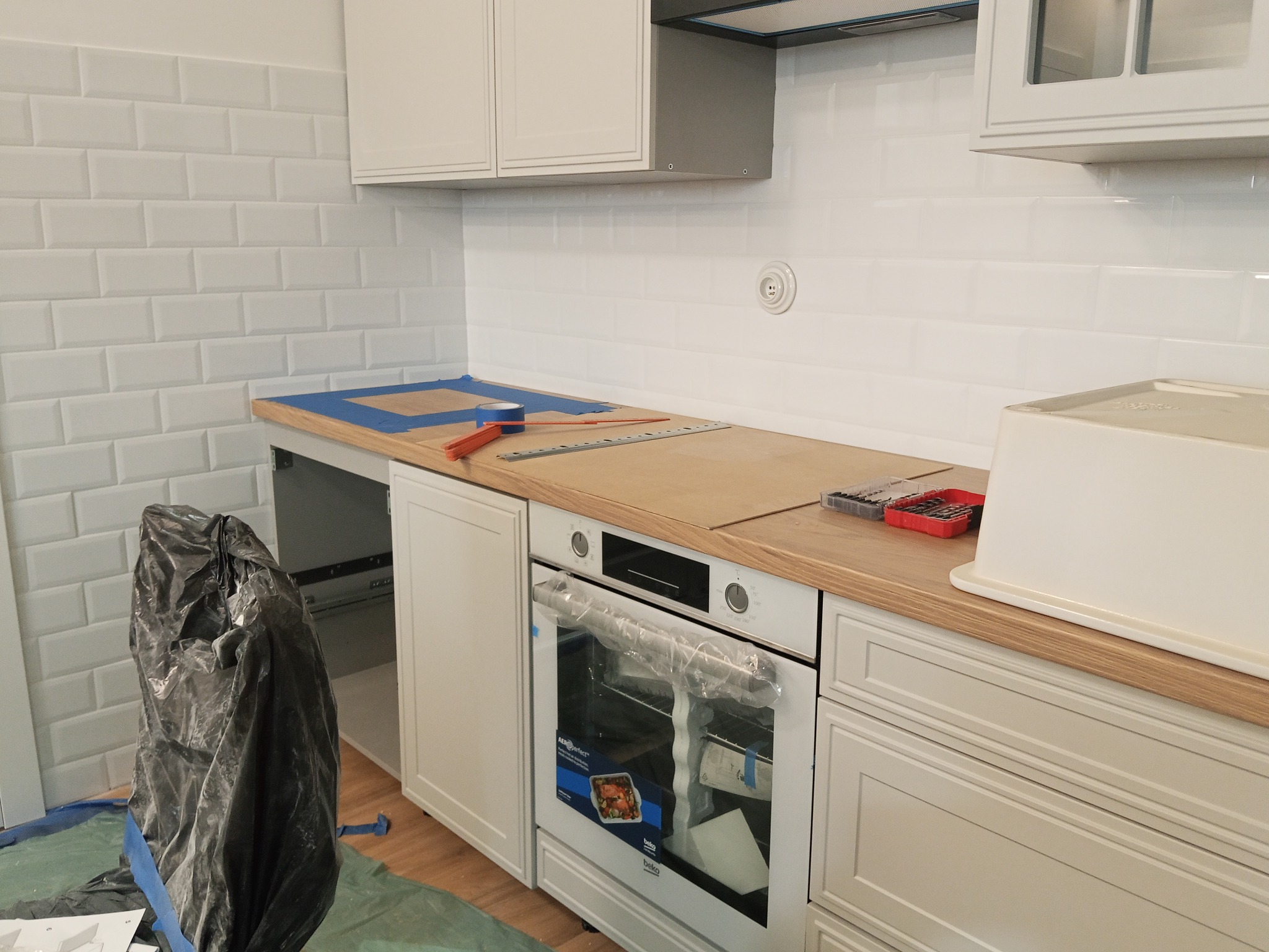Biała kuchnia w trakcie montażu: zabudowa z piekarnikiem, drewniany blat, białe płytki na ścianie. Widoczne narzędzia i folia ochronna na sprzęcie.