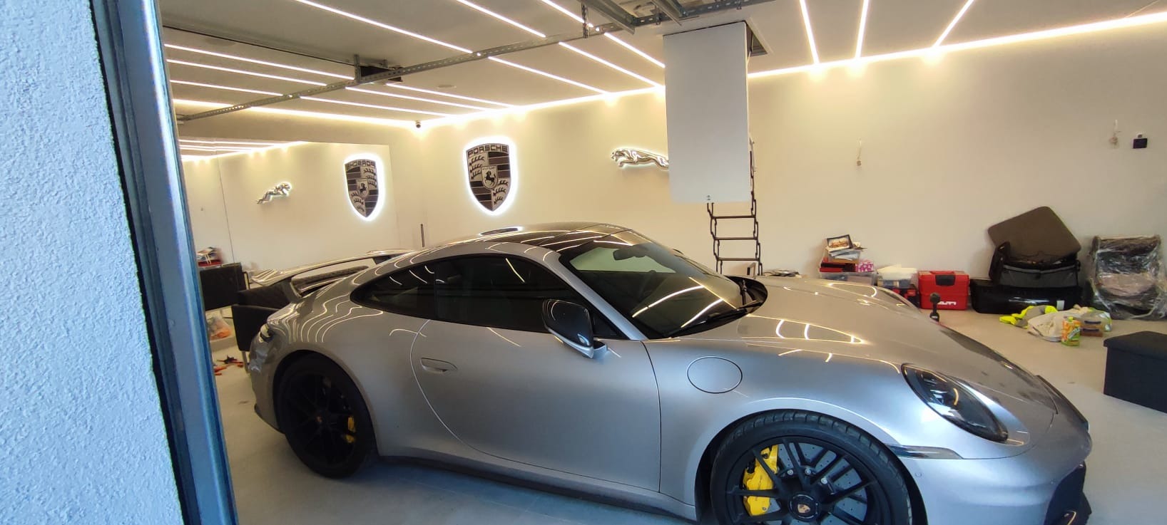 Nowoczesny garaż z oświetleniem LED i srebrnym Porsche. Na ścianie podświetlane logo Porsche i Jaguar. Schody strychowe opuszczone. Widoczne akcesoria samochodowe.