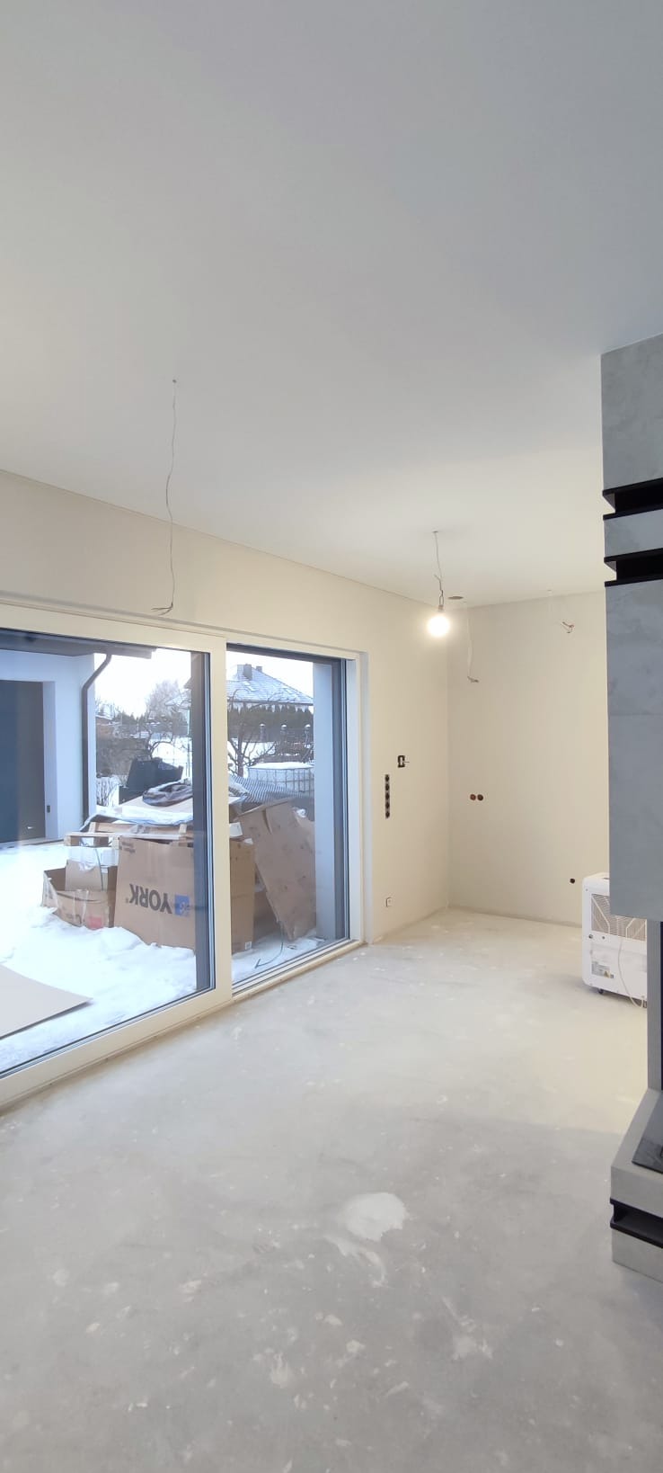 Wnętrze w trakcie remontu: surowe ściany, wylewka podłogowa, okno z widokiem na zimowy krajobraz. Widoczne puszki elektryczne i kartony.