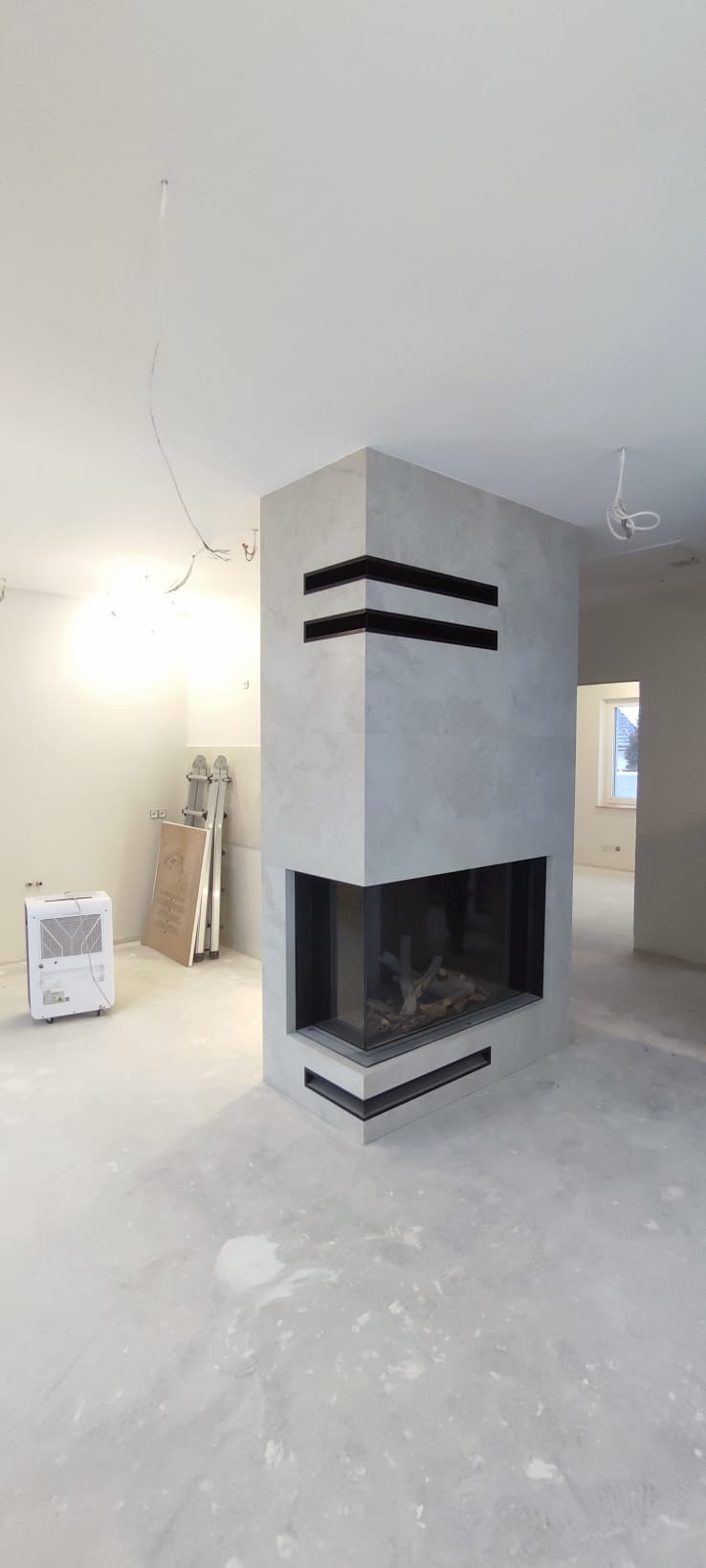 Nowoczesny kominek obudowany szarym kamieniem z czarnymi akcentami w surowym wnętrzu. Widoczne niedokończone prace wykończeniowe i drabina.