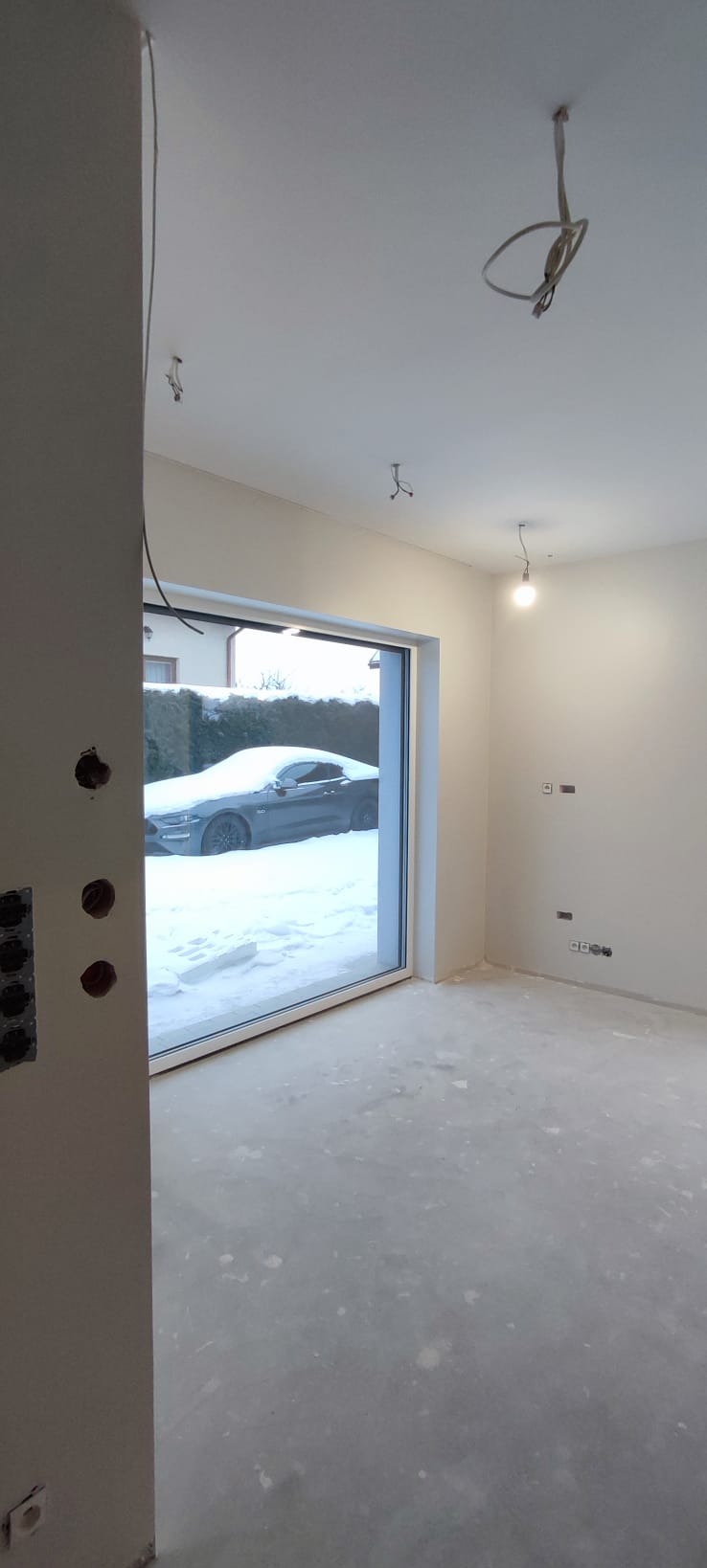 Wnętrze w trakcie prac wykończeniowych: surowa podłoga, ściany przygotowane do malowania, widoczne instalacje elektryczne. Za oknem ośnieżony samochód.