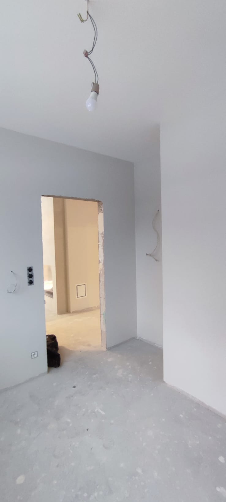 Wnętrze w trakcie prac wykończeniowych: świeżo pomalowane ściany na szaro, surowa podłoga, widoczne kable i gniazdka. W tle otwarte przejście do innego pomieszczenia.