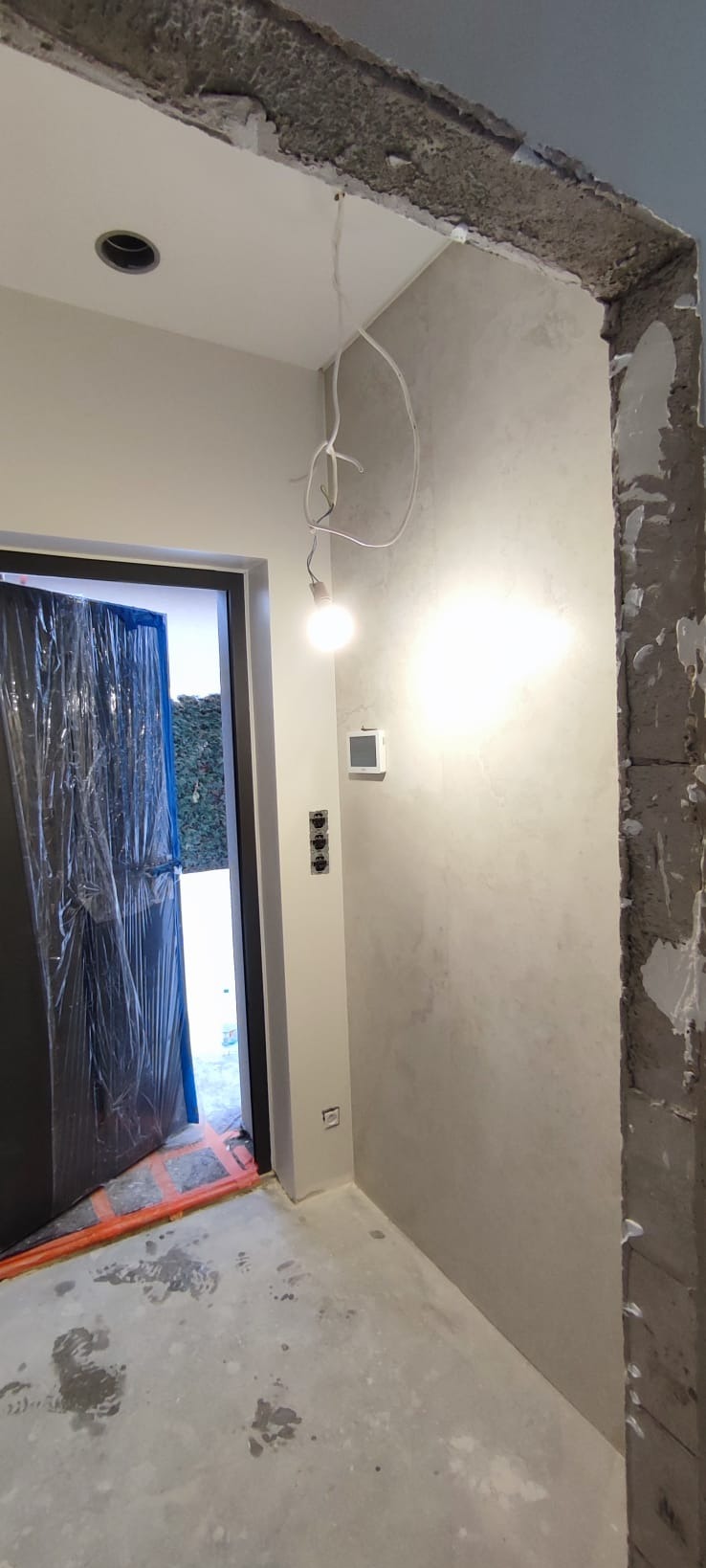 Wnętrze w trakcie wykończenia: surowa ściana z widoczną fakturą, zabezpieczone folią drzwi, wystające kable z żarówką. Widoczne gniazdka i włączniki.