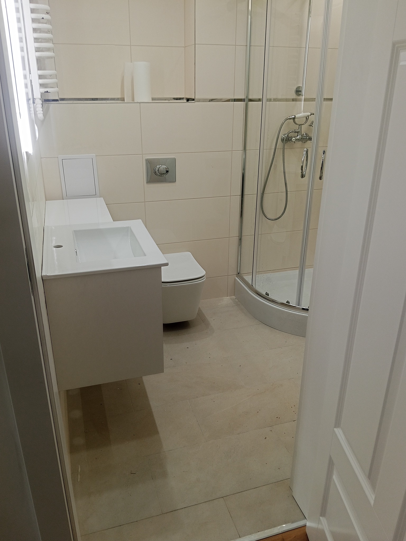 Jasna łazienka po remoncie: kabina prysznicowa, umywalka z szafką, toaleta. Beżowe płytki na ścianach i podłodze. Widok zza uchylonych drzwi.