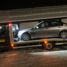 EMJ Jakub Oleśkiewicz - Srebrny samochód osobowy na platformie lawety w zimowej scenerii nocą. Widoczny napis 'Pomoc Drogowa' na pojeździe. Śnieg na karoserii i wokół.