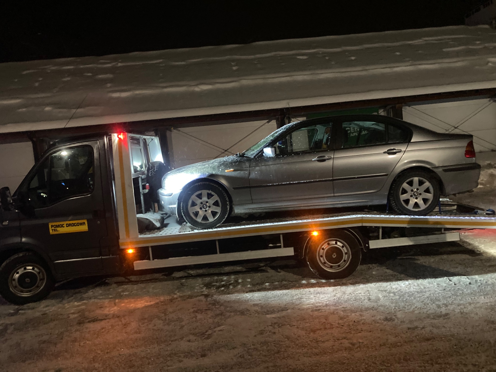 Srebrny samochód osobowy na platformie lawety w zimowej scenerii nocą. Widoczny napis 'Pomoc Drogowa' na pojeździe. Śnieg na karoserii i wokół.