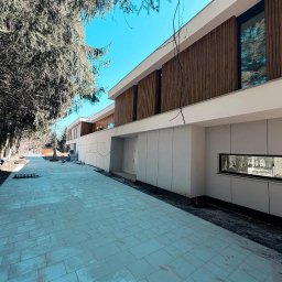 SUBSIDIA SPÓŁKA Z OGRANICZONĄ ODPOWIEDZIALNOŚCIĄ - Nowoczesny dom z elewacją z paneli drewnianych i jasnoszarych płyt. Widok z zewnątrz, z brukowanym podjazdem i drzewami w tle. Minimalistyczny design z długim, poziomym oknem.