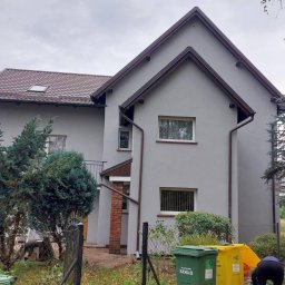 SUBSIDIA SPÓŁKA Z OGRANICZONĄ ODPOWIEDZIALNOŚCIĄ - Dom jednorodzinny po remoncie elewacji, z nowymi oknami i rynnami w kolorze brązowym. Widoczne kosze na śmieci i fragment osoby w ogrodzie.