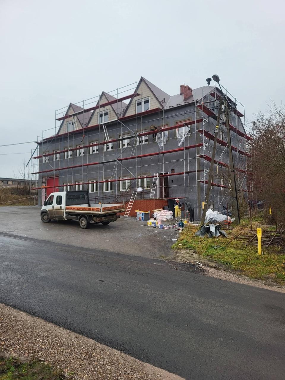 Ocieplanie budynku wielorodzinnego z użyciem rusztowań. Widoczny samochód dostawczy i pracownicy przygotowujący materiały. Szara, pochmurna pogoda.