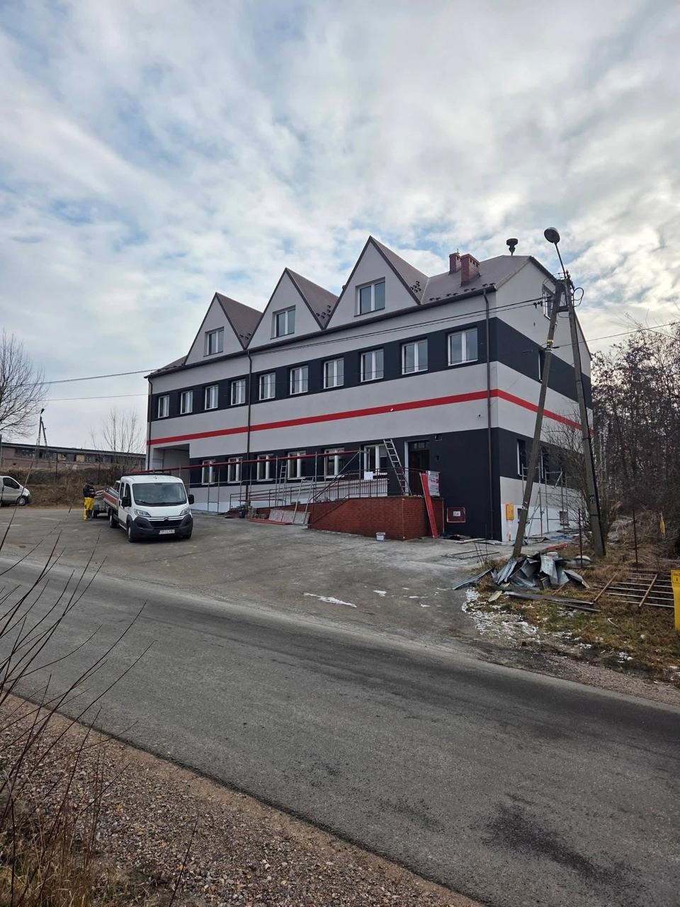 Trzypiętrowy budynek po renowacji elewacji z szarymi i czarnymi pasami, czerwoną linią i trzema szczytowymi dachami. Na pierwszym planie ulica i samochód dostawczy.