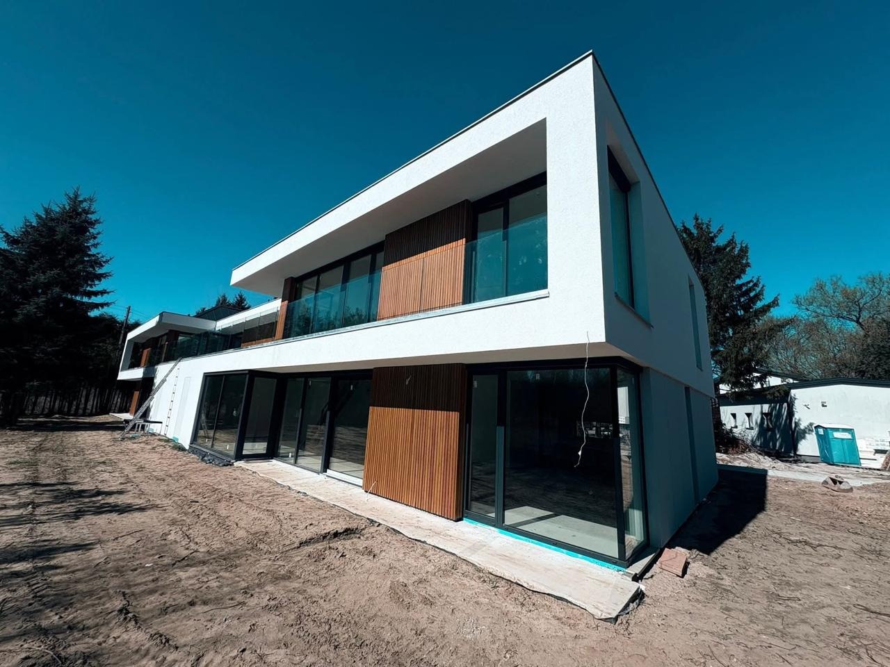 Nowoczesny, dwukondygnacyjny dom z białymi ścianami i drewnianą elewacją, duże okna, minimalistyczny design, otoczony drzewami, w słoneczny dzień.