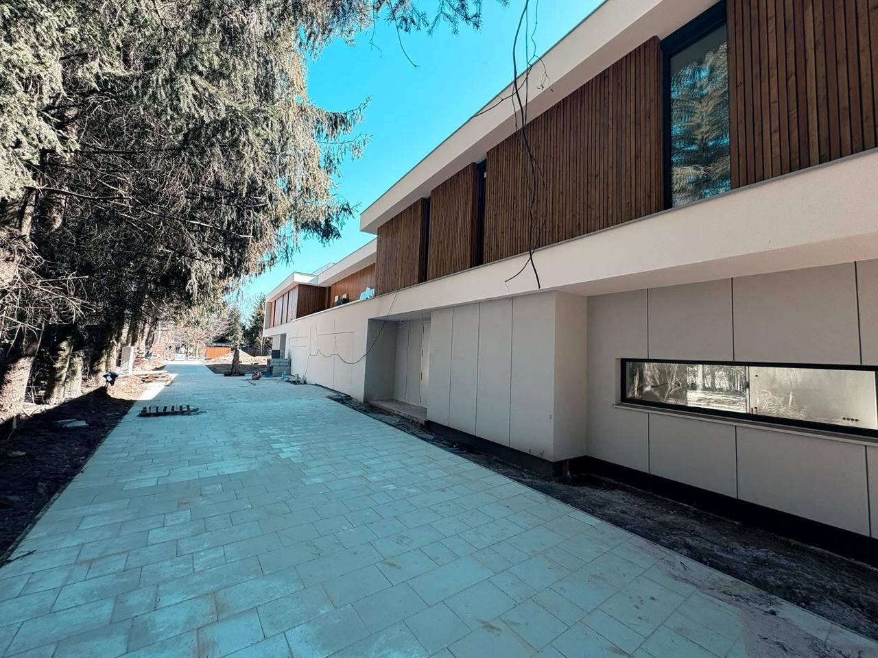 Nowoczesny dom z elewacją z paneli drewnianych i jasnoszarych płyt. Widok z zewnątrz, z brukowanym podjazdem i drzewami w tle. Minimalistyczny design z długim, poziomym oknem.