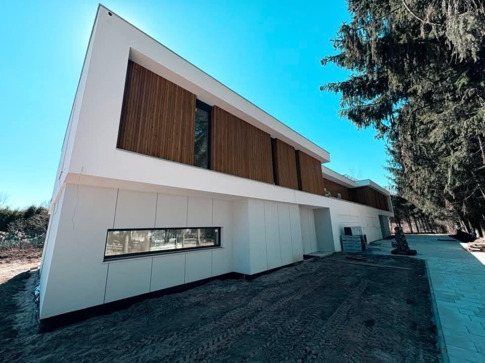 Nowoczesny dom z białą elewacją i drewnianymi panelami, widok z dołu, błękitne niebo w tle. Minimalistyczny design, surowe wykończenie, teren wokół domu przygotowany pod zagospodarowanie.